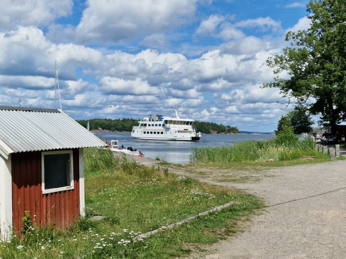 Ett fartyg nära strandkanten, molnig himmel, grönska, mindre byggnad, fridfull sommardag vid vattnet.