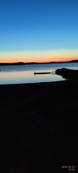 En brygga vid en sjö under en färgstark solnedgång med klarblå himmel övergående i orange och mörkblått.