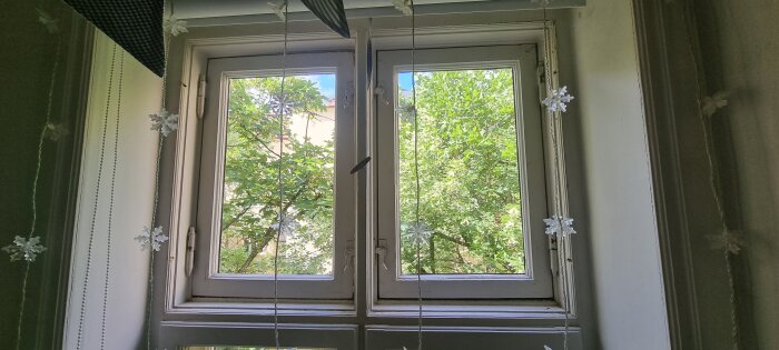 Traditionellt fönster med öppna fönsterluckor, vita snöflingedekorationer och utsikt över gröna träd under sommaren.