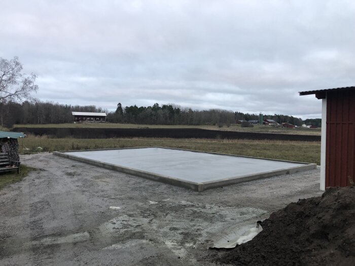 Nybyggd betongplatta på landsbygd, gråmulet väder, öde landskap, jordbruksmark i bakgrunden, lite snö.