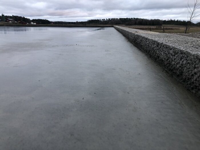 Betonkaj vid vattnet, isbelagd sjö, disig himmel, skog i bakgrunden, molnig vinterdag.