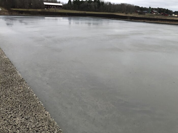 Isfrusen sjö vid landsbygd, grå himmel, grusväg förgrunden, träbyggnader i bakgrunden, lugnt vatten, vinter eller tidig vår.