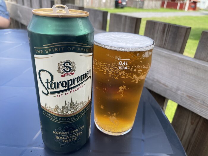 En Staropramen ölburk och ett glas öl utomhus på ett träbord.