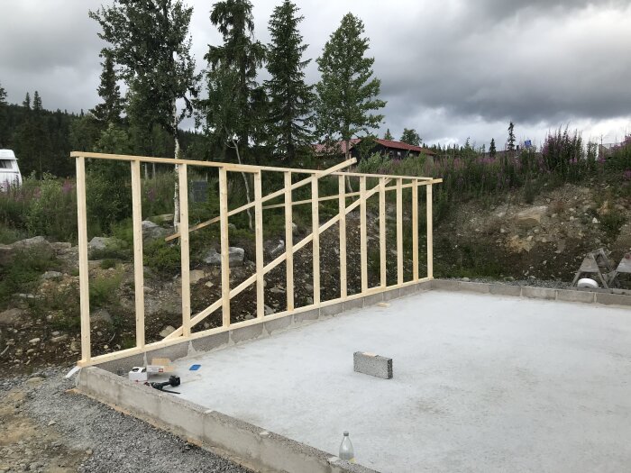 Byggnadsställning i trä på betonggrund med natur och molnig himmel i bakgrunden. Verktyg synliga.