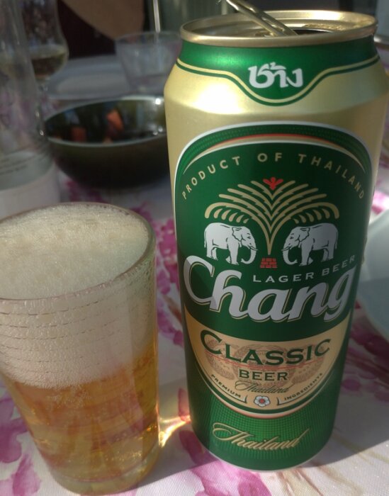 Chang ölburk med glas, skummande öl, utomhus, ljus bakgrund, matbord, "Product of Thailand"-text.