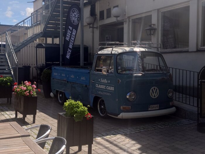 En blå VW-buss framför en trappa och en byggnad, omgiven av blomlådor och tomma uteserveringsbord.