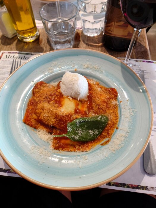 En lasagneportion med smält ost och basilikablad, serverad med en kula ricotta, på en turkos tallrik.