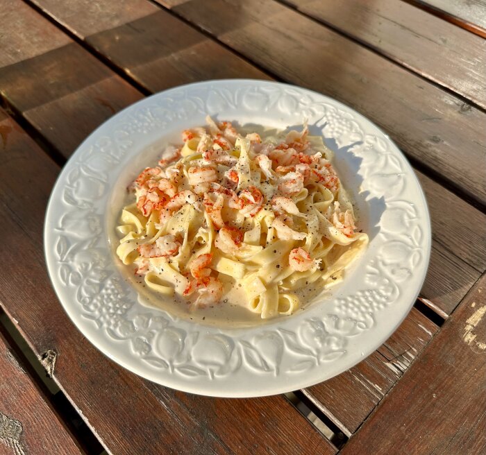 Tallrik med pasta och räkor på träbord utomhus i solsken.