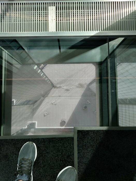 Glastaksgolv, person ser ned, hög höjd, urban miljö, sneakers synliga, perspektivförvrängning, arkitektoniska detaljer, vertikal utsikt.