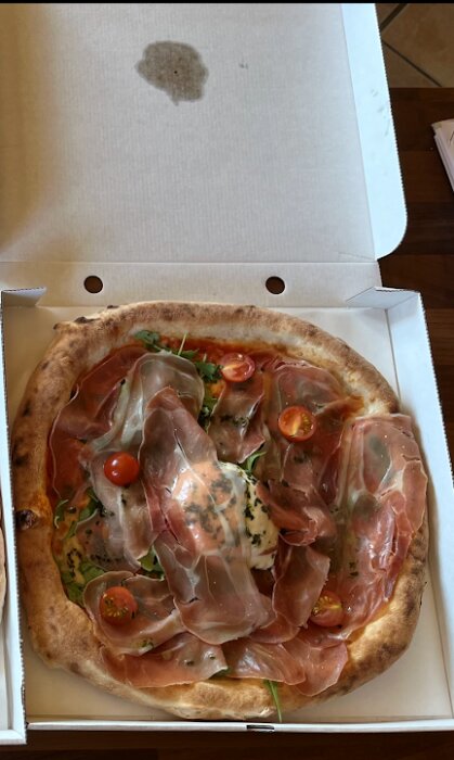 En pizza med prosciutto, tomater och rucola i en kartong. Oljefläck på locket.