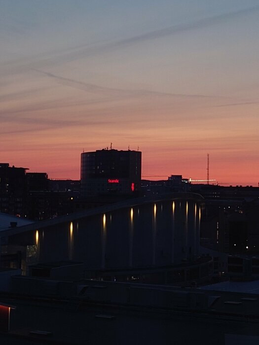 Stadsilhuett vid solnedgång, himlen tonar från blått till rosa, ljus från byggnader, rofyllt kvällsljus.