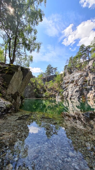 Klippiga bergssluttningar, gröna träd, klarblå himmel, spegelblank grön sjö, naturskönt område, reflektion i vatten.