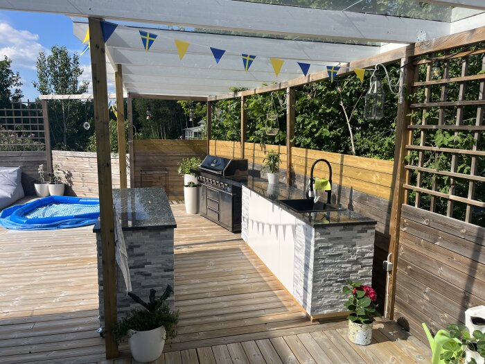 Uteplats med sommarkök, grill, diskho, vimplar, loungemöbler och uppblåsbar pool. Trägolv, grönska, avskärmningar, soldränkt.