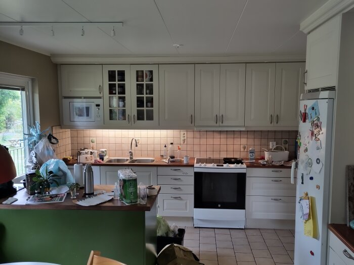 Ett kök med vita skåp, hushållsapparater och diverse föremål på diskbänken och bordet; använd och bebott rum.