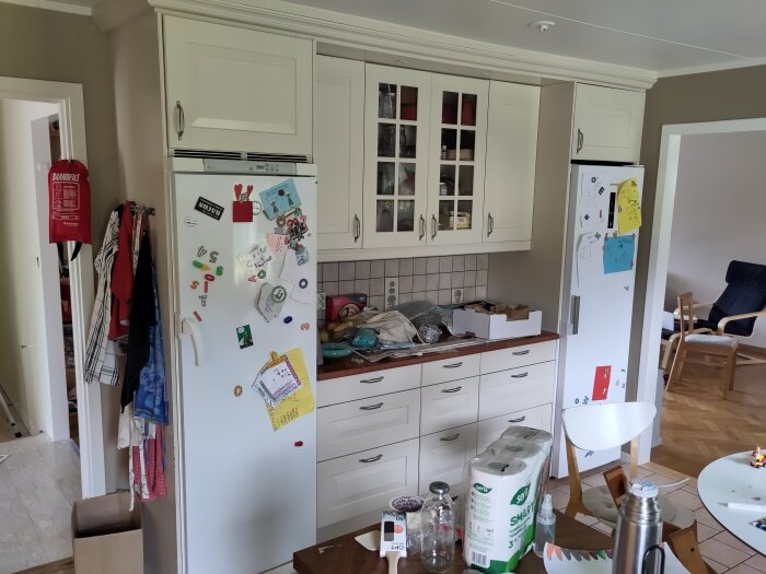 Ett kök med vit skåpinredning, kylskåp täckt av magneter och teckningar, oordnad bänkyta.