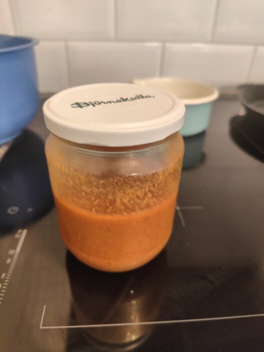 Ett glas med orange innehåll och vit lock på en spishäll, köksbakgrund.