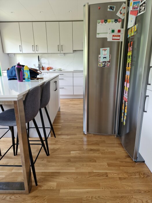 Modernt kök med vita skåp, rostfri kyl, trägolv, grå barstolar och dekorationer på köksön.