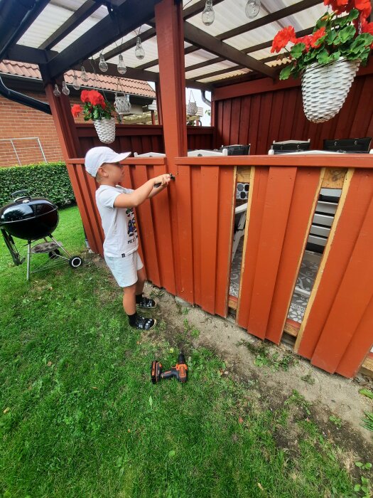 Pojke skruvar på en träspaljé under pergola utomhus, leksaksbil och trädgårdsverktyg synliga på gräset.