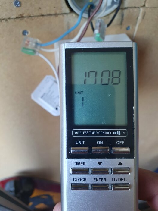 Handhållen digital timer visar kl. 17:08, bakgrund med oskarpa elektroniska komponenter och vägg.