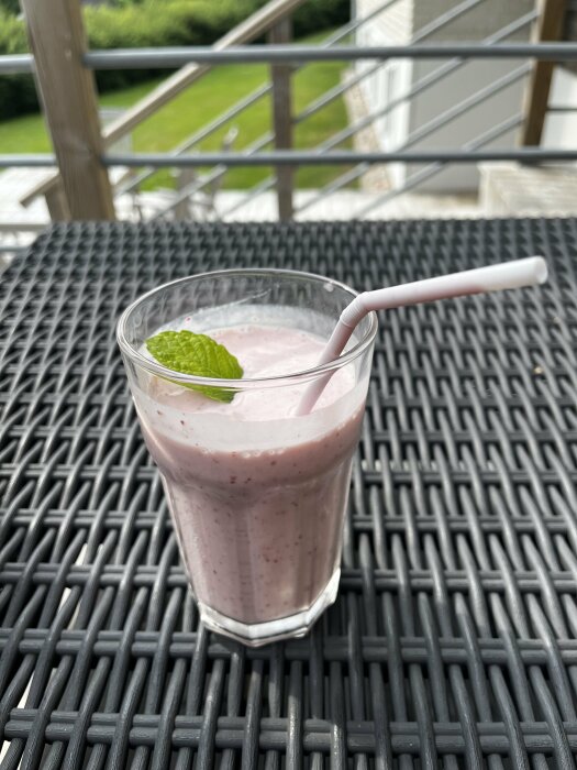 Ett glas med rosa smoothie och sugrör på flätat bord, utomhus, med mintblad på toppen.