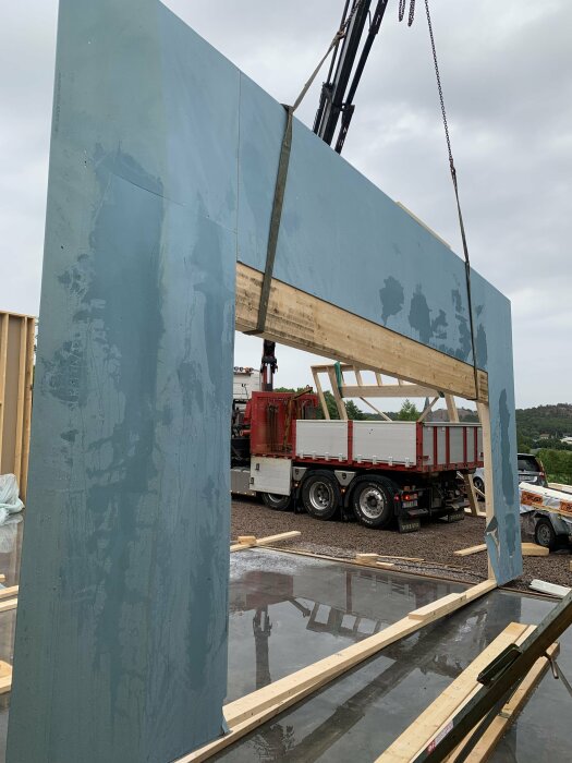 Lyftkran höjer stor betongpanel över byggplats med lastbil och spegelbild i vattenpöl.