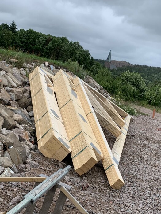 Staplade träbjälkar på stenig mark, skog och kyrktorn i bakgrunden, molnig himmel, byggnation eller konstruktionsplats.