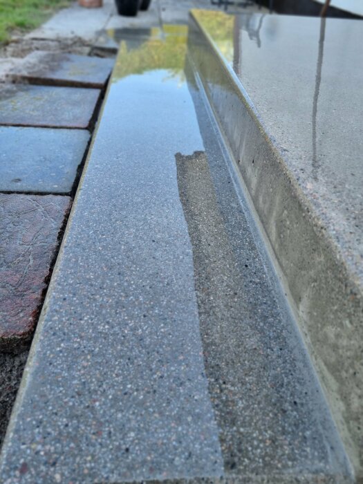 Reflektion av himmel och byggnad i vattenpöl på grå trottoarkant och gatsten.