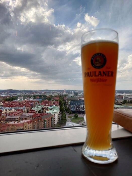 Ölglas med Paulaner-logo framför stadsutsikt och molnig himmel, fotograferat från högt perspektiv.