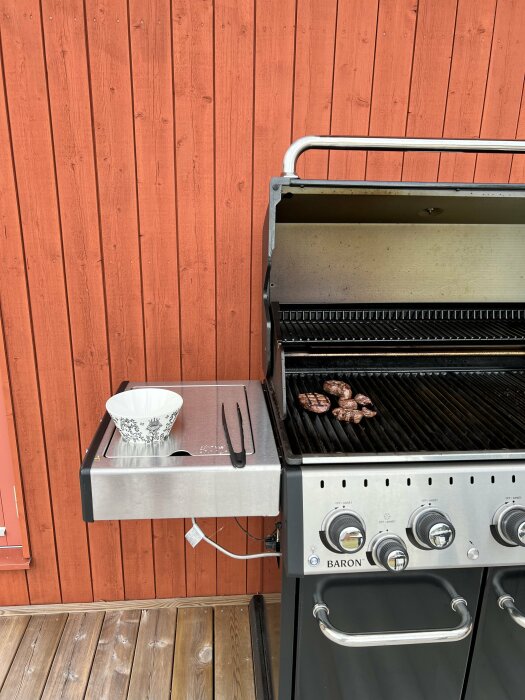 Gasolgrill på veranda, grönsaker på värmning, tänger, träslevar, skål på sidobord, röd trävägg, trägolv.