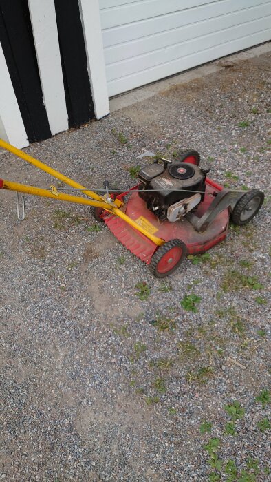 Gammal gräsklippare på grus uppfart nära garageport, röd, gul, lite övervuxet gräs.