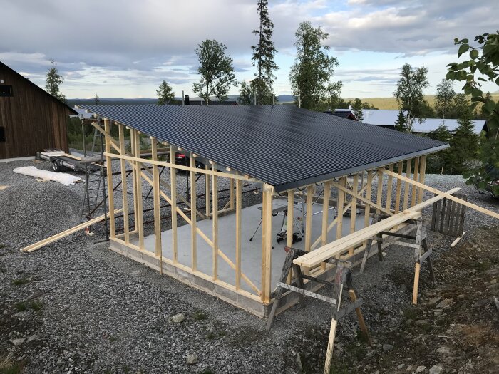 Ett pågående bygge av en träkonstruktion med svart plåttak, utomhus vid skymningen med träd och utsikt.