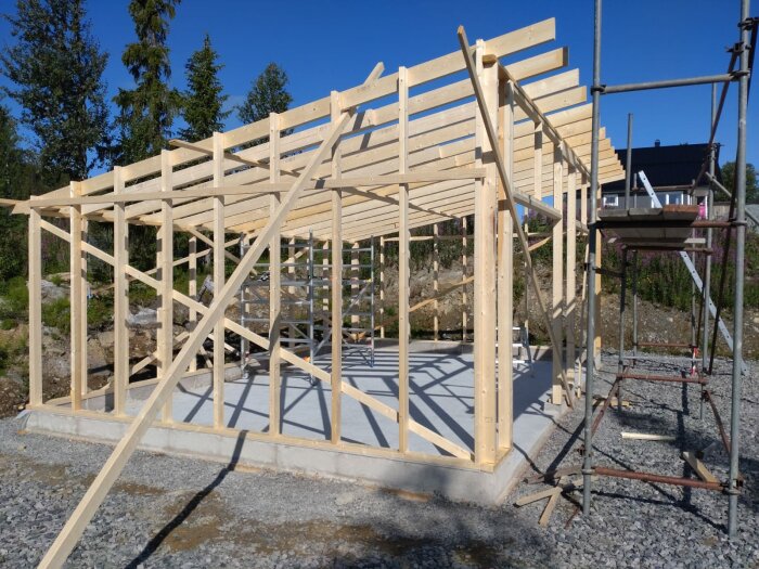 Trästomme för byggnad under konstruktion, med blå himmel och ställning, på en betonggrund.