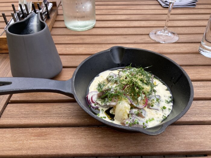 Maträtt i stekpanna på träbord, gräddig sås, rödlök, dill, utomhus, bestickhållare, vattenglas, tomma glasfot.