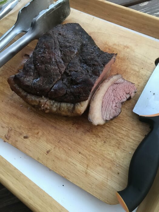 Ett tillagat köttstycke på skärbräda med kniv och grilltång, delvis uppdelat, rosa insida.