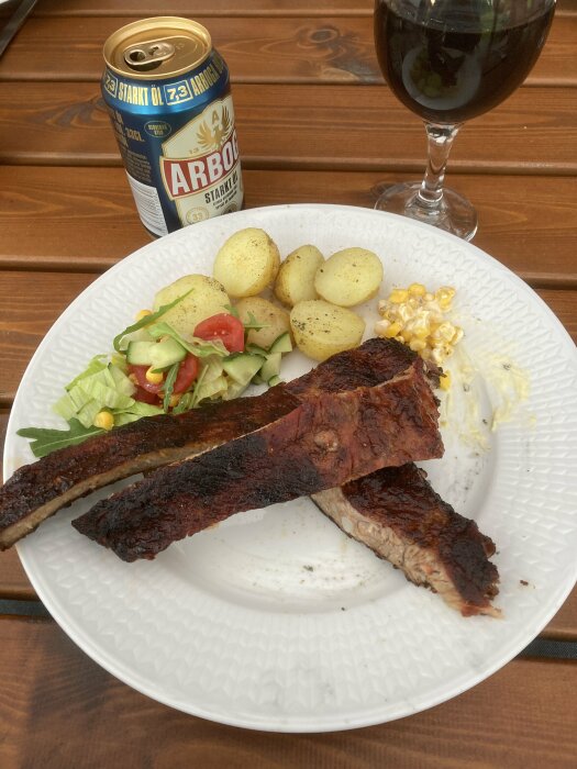 Grillade revbensspjäll, kokt potatis, sallad, öl och ett glas mörk dryck, serverade utomhus.