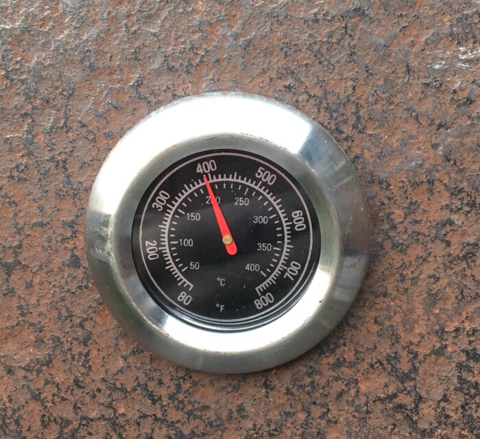 Termometer i metall på brun bakgrund visar cirka 210 grader Fahrenheit och ungefär 100 grader Celsius.