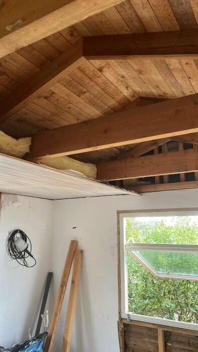Renovering pågår, öppet tak med exponerade träbjälkar, och isolering, vägg och fönster synliga.
