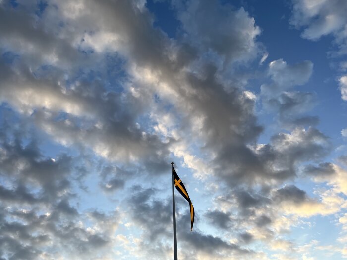 En flaggstång med Sveriges flagga mot en dramatisk himmel fylld med blått och moln vid solnedgång eller soluppgång.