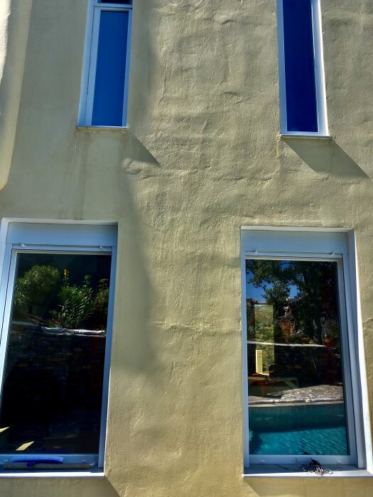Soligt hus, beige vägg, blå fönsterkarmar, spegling av trädgård och pool i fönstret.