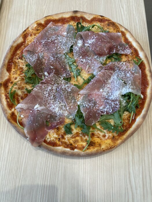 En pizza med prosciutto, rucola och riven ost på ett träbord.