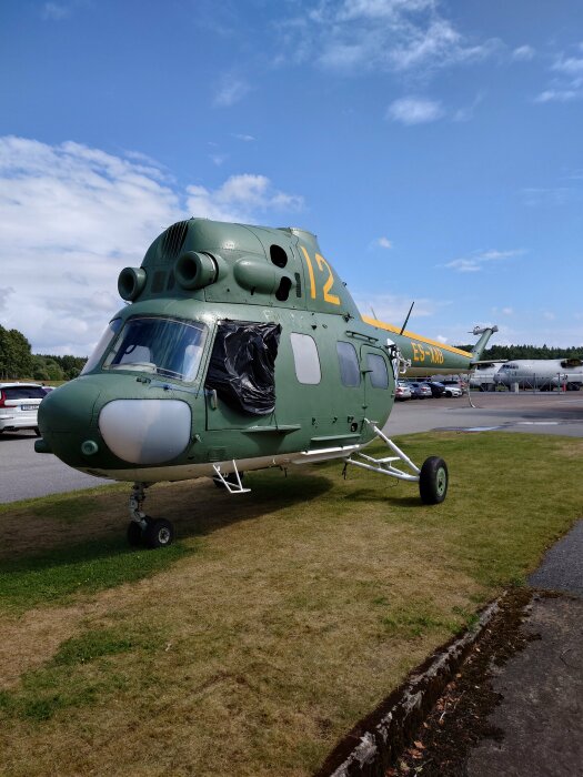 Grön militärhelikopter parkerad på gräs med täckt fönster och klarblå himmel.