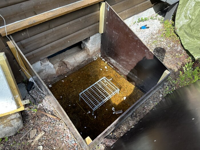 Vattenfylld grund vid byggnad, trådhylla i vatten, elektrisk sladd, trävägg, grus, tarp, utomhus.