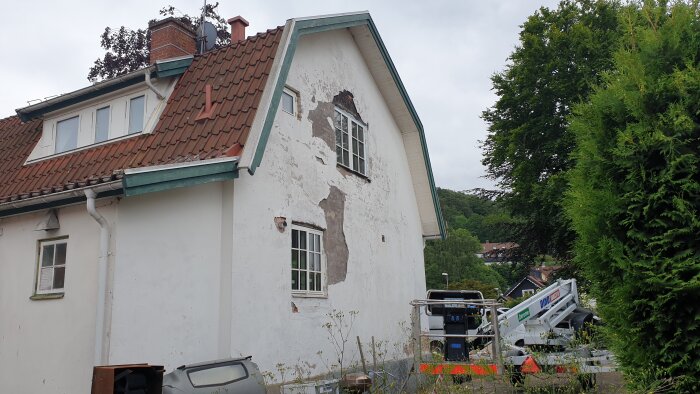 Ett vitputsat hus med sliten fasad, takpannor och grönska runtomkring. Arbetsfordon och skyltar syns framför.