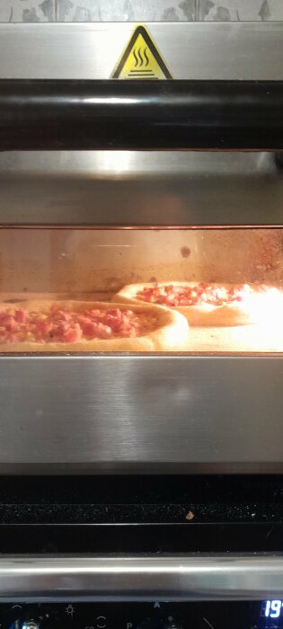 Två pizzor bakas i en industriell ugn, synliga genom glasdörren. Varningstecken för het yta ovanför.