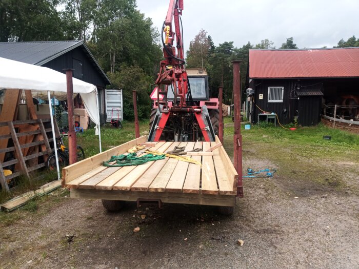 Lastbilsflak med träplankor, röd kranarm, lantgårdsmiljö, skjul, hötält, grönt gräs, cykel och verktyg synliga.