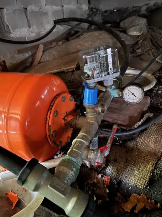 Vattenpump och tryckmätare i rörigt utrymme med bråte och ledningar.