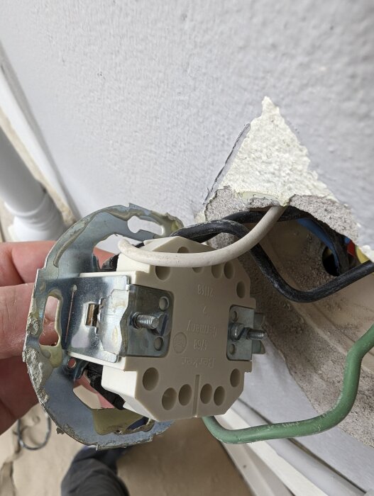 En person håller i en demonterad elektrisk vägguttag under installation eller reparation. Kablar och väggstruktur syns.