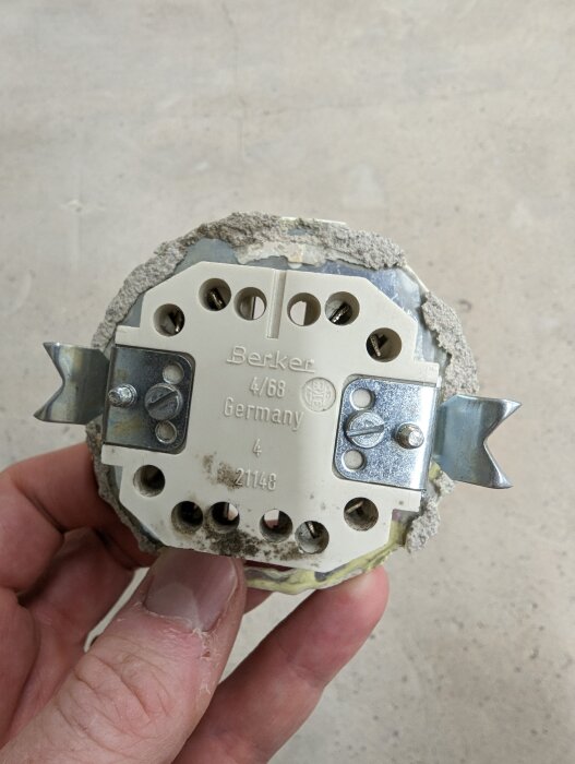 Hand håller en gammal elektrisk installationsdosa med Berker märke, betongrester och skruvterminaler, från Tyskland.