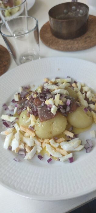 Rätten potatissill med lök, kokt ägg, på vit tallrik, oskarp bakgrund med glas, kopparunderlägg.