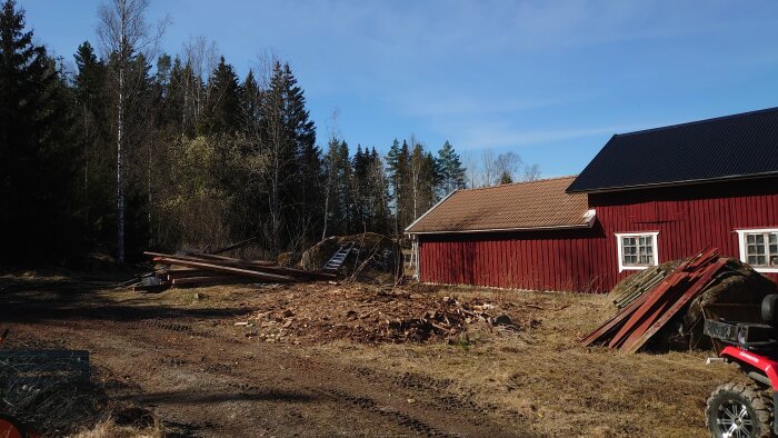 En röd stuga, skog, himmel, raserad byggnad och en fyrhjuling på en lantlig plats.